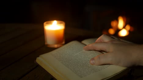Ein-Mann,-Der-Ein-Altes-Antikes-Buch-An-Einem-Gemütlichen-Feuer-Liest-Und-Von-Kerzenlicht-Aus-Nächster-Nähe-Beleuchtet-Wird