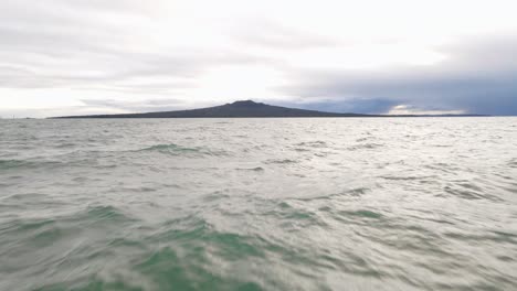 Fast-and-low-aerial-approach-from-a-beach-towards-Rangitoto-island-during-a-cloudy-sunrise