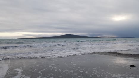 Small-waves-rolling-ashore-with-the-magnificent-Rangitoto-volcano-in-the-background-and-dramatic-clouds-above