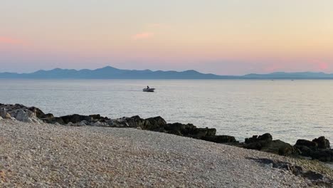 Un-Pescador-Navega-En-Un-Bote-Al-Atardecer,-Vista-Desde-La-Playa,-Toma-De-Mano,-Croacia