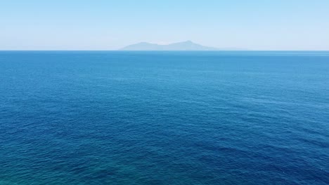 Aerial-drone-rising-over-vast-blue-ocean-and-rugged,-remote-tropical-Atauro-Island-in-the-distance-as-seen-from-capital-Dili,-Timor-Leste,-Southeast-Asia