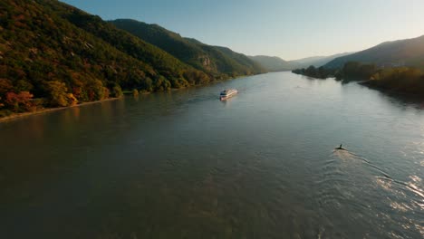 Danube-boat-decorated-with-multiple-flags-approaching-FPV-drone,-passing-by-at-close-proximity-and-making-waves-reflecting-the-beautiful-sunset-in-wachau-valley