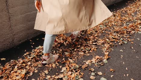 Woman-is-walking-through-fallen-brown-tree-leaves-and-near-the-concrete-wall,-autumn-atmosphere