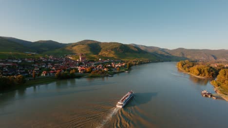 Slow-ascending-FPV-drone-shot-of-a-danube-boat-passing-Weissenkirchen-town-in-wachau-valley-dureing-sunset-time-in-autumn