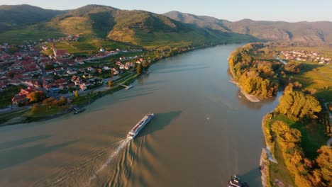 Danube-boat-slowly-going-down-the-danube-river-passing-the-town-Weissenkirchen,-while-making-small-waves-reflecting-the-light-of-the-beautiful-autumn-evening-sun