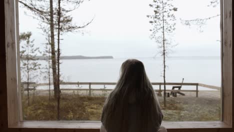 Girl-is-watching-the-quiet-lake-landscape-outside-of-the-window-of-her-home