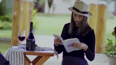 Fashionable-girl-wearing-fedora-opening-book-by-a-glass-of-red-wine-and-bottle-at-a-winery