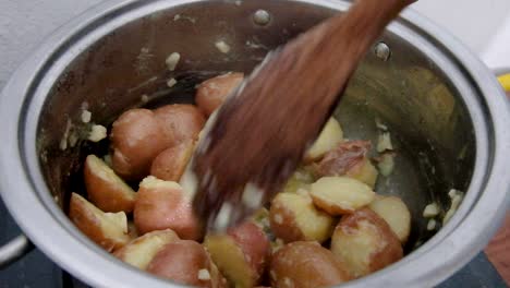 Kochen-Und-Rühren-Neuer-Kartoffeln-Mit-Einem-Großen-Holzlöffel,-Der-In-Knoblauchbutter-In-Einem-Edelstahltopf-Köchelt,-Nahaufnahme-Von-Köstlichen-Kartoffeln-Mit-Roter-Haut