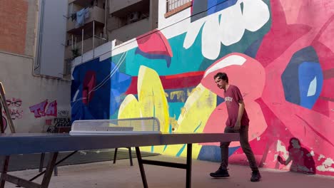 20-year-old-boy-playing-ping-pong-in-a-street-tennins-table-with-his-father-with-a-colorful-flower-graffiti-at-his-back