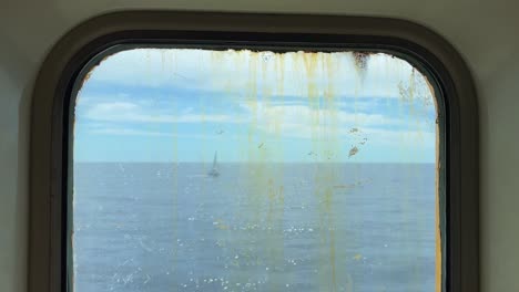 Sailboat-sailing-on-calm-sea-as-seen-through-dirty-and-rusty-window-of-navigating-old-ferry