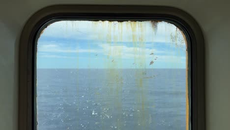 Sailboat-sailing-on-calm-sea-water-as-seen-through-dirty-and-rusty-window-of-navigating-old-ferry