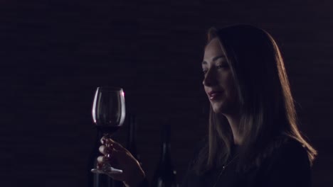 Brunette-woman-raising-red-wine-glass-in-a-dark-ambient-romantic-studio-room,-looking-directly-into-camera