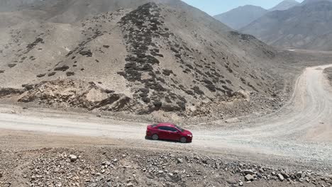 Aerial-panning-shot-of-a-road-trip-with-a-red-car-on-a-dry-dusty-country-road-with-stones-along-the-hills-to-the-holy-city-of-Caral-in-peru-on-a-sunny-day