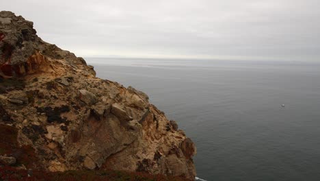 Panning-shot-of-jagged,-high-rocky-cliff-with-a-lone-boat-sailing-on-a-gray-ocean-below