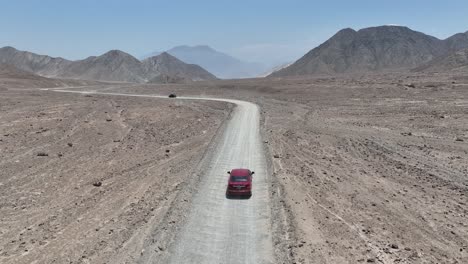 Toma-Aérea-De-Un-Vehículo-Rojo-Y-Negro-Conduciendo-Por-Una-Carretera-Polvorienta-En-El-Desierto-De-Perú-Frente-A-La-Ciudad-Santa-De-Caral-Con-Colinas,-Montañas-Y-Rocas-En-Un-Día-Soleado