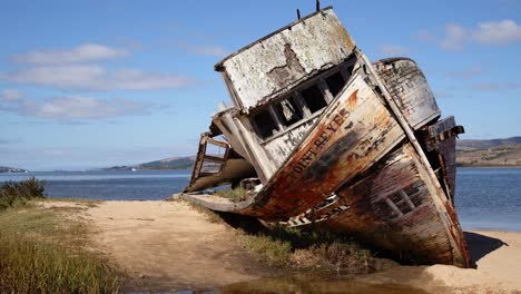 Panning-shot-of-rotting,-shipwrecked-Point-Reyes-boat-washed-up-on-a-sunny-Tomales-Bay-shore