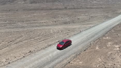 Aerial-panning-shot-of-a-moving-red-vehicle-in-the-desert-of-peru-in-front-of-the-holy-city-of-caral-on-a-dusty-highway-overlooking-the-dried-landscape-with-hills-in-the-background