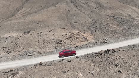 Aerial-panning-shot-of-a-red-vehicle-while-traveling-on-a-dusty-and-dry-highway-in-peru-along-the-hills-and-stones-on-the-side-near-the-holy-city-of-caral-on-a-sunny-day