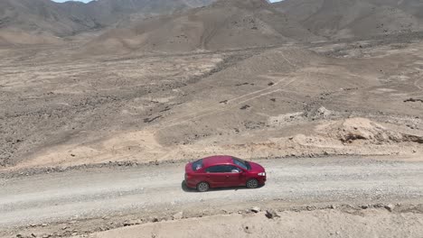 Aerial-panning-shot-of-a-red-vehicle-driving-in-the-desert-of-peru-towards-the-holy-city-of-caral-on-a-dusty-highway-with-hills,-stones-and-mountains-in-the-background-on-a-sunny-day