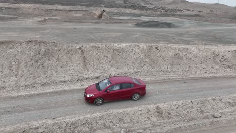 Aerial-panning-shot-of-a-red-vehicle-driving-along-a-dusty-and-stony-road-in-the-desert-along-stone-hills-in-front-of-the-holy-city-of-Caral