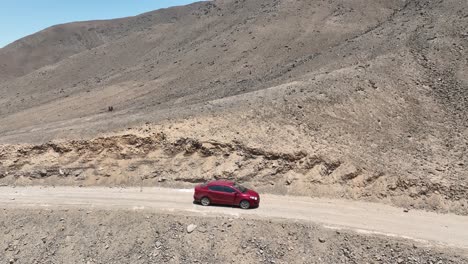 Aerial-panning-shot-of-a-red-moving-car-in-the-wasteland-of-peru-on-a-dusty-dry-desert-road-along-the-hills-towards-the-holy-city-of-caral