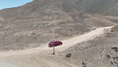 Aerial-panning-shot-of-a-red-moving-vehicle-during-a-short-trip-in-the-desert-country-of-peru-on-a-dusty-highway-with-hills,-stones-and-mountains-on-a-cloudless-sunny-day