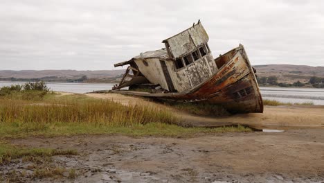 Panning-shot-of-rotting,-shipwrecked-Point-Reyes-boat-washed-up-on-a-muddy-Tomales-Bay-shore