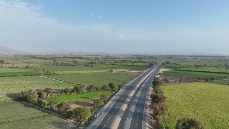 Extreme-long-aerial-shot-of-vehicles-crossing-four-lane-Panamericana-Norte-highway-surrounded-by-mountains-in-Peru-through-farming-fields-on-both-corners
