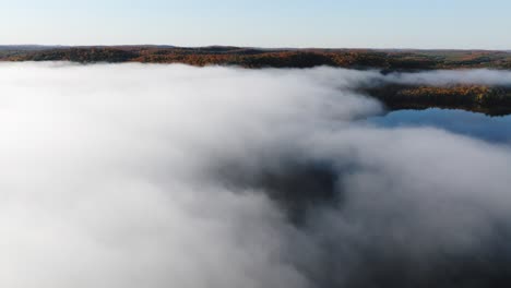 Aerial-close-up-of-fog-above-body-of-water,-colourful-Autumn-trees-in-the-background,-during-cool,-bright,-sunny-day