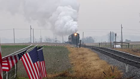 View-of-a-Steam-Passenger-Train-Approaching-Blowing-Smoke-and-Steam-With-American-Flags-Waving-in-the-Wind-on-the-Fence-on-a-Cloudy-Day