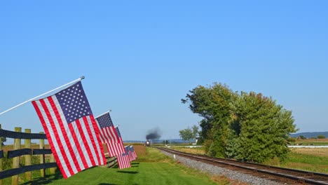 View-of-a-Steam-Passenger-Train-Approaching-Blowing-Smoke-and-Steam-With-American-Flags-Waving-in-the-Wind-on-the-Fence