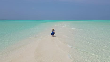 Young-girl-wearing-a-long-blue-dress-running-barefoot-on-a-surreal-white-sandbank-with-turquoise-waters-of-Indian-ocean,-Maldives-drone-shot-on-a-clear-sunny-day
