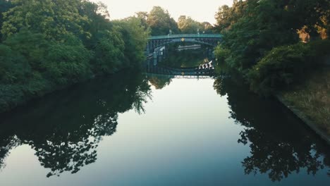 Drone-aerial-river-in-a-park-during-sunrise-with-a-bridge-reflecting-on-the-water