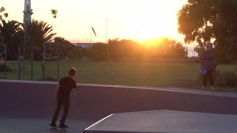 Slow-motion-shot-of-boy-dropping-in-at-a-skate-park-skateboarding-during-sunset