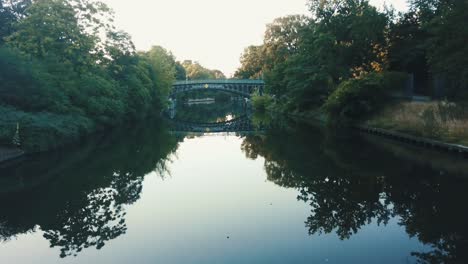 Drone-aerial-moving-backwards-over-river-in-a-park-during-sunrise-with-a-bridge-reflecting-on-the-water