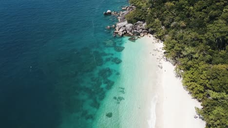 Drone-aerial-over-tropical-blue-clear-water-and-white-sandy-beach-with-a-foresty-island