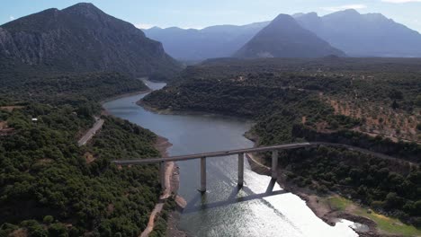 Aerial-View-Of-Strada-Provinciale-38-Road-Crossing-River-Lago-del-Cedrino-With-Mountain-Landscape-In-The-Background