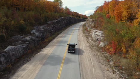 Country-road-with-pick-up-truck-leading-through-autumn-forest-and-rocky-cliffs-sides