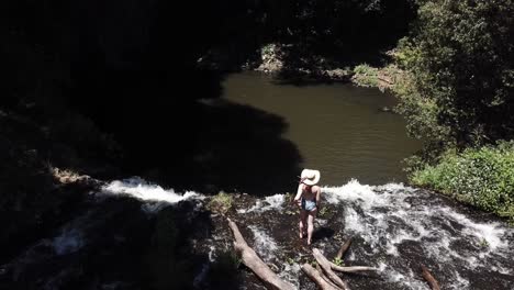 Drone-aerial-in-nature-with-a-woman-walking-on-the-ledge-of-a-waterfall-exploring-with-the-camera-panning-down