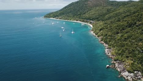 Drone-aerial-pan-up-over-tropical-clear-blue-water-foresty-island-with-boats