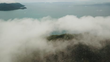 Drone-aerial-above-clouds-on-tropical-forest-island-panning-down-to-reveal-trees-and-trail
