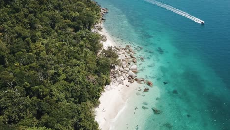 Drone-aerial-over-tropical-blue-clear-water-and-white-sandy-beach-with-a-foresty-island-with-a-speed-boat