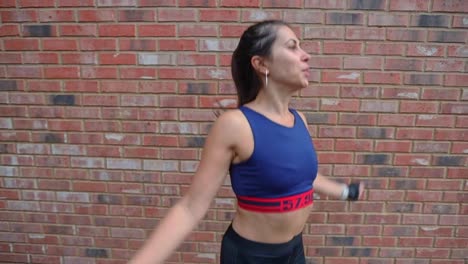 Sporty-active-Hispanic-female-doing-jumping-jacks-warm-up-in-front-of-brick-wall