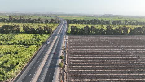 Filmische-Luftaufnahme-Von-Oben-Nach-Unten,-Die-Einem-Auto-Auf-Der-Mehrspurigen-Peruanischen-Autobahn-Panamericana-Norte-Zwischen-Ausgedehnten-Ackerflächen-An-Beiden-Ecken-Folgt