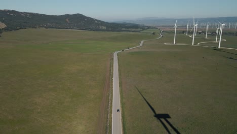 High-angle-view-following-vehicle-travelling-long-road-through-sustainable-wind-turbine-farm