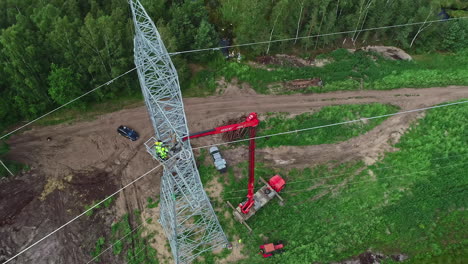 Aerial-top-down-shot-of-industrial-worker-lifting-by-crane-repairing-electricity-pylon-in-woodland