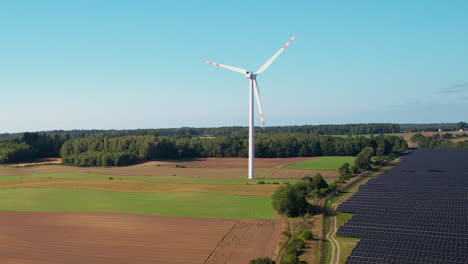 Solar-Panels-With-Wind-Turbine-On-Countryside-Landscape-In-Summer---drone-shot