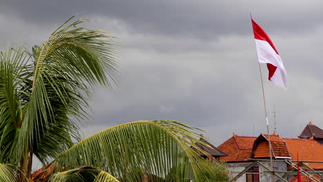 Merah-Putih,-Indonesische-Flagge,-Die-Gegen-Stürmische-Wolken-Mit-Palmen-Und-Traditionellen-Dächern-Weht
