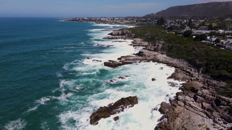 Cinematic-drone-flight-over-rugged-Hermanus-coastline-with-waves-hammering-rocks---historic-fishing-village-on-Cape-Whale-Coast