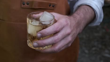 Man-in-leather-apron-holds-a-whiskey-glass-with-ice